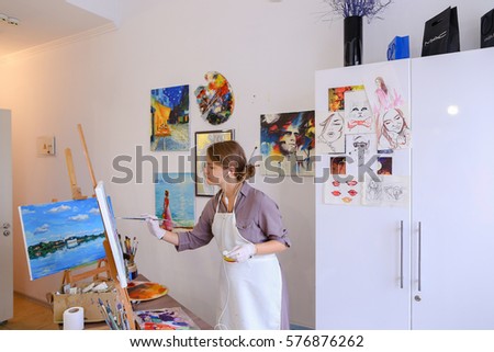Artist attractive woman of European appearance writes oil painting, enjoying things what love , paint picture for sales and order, listening to music on headphones through phone, stands and works in