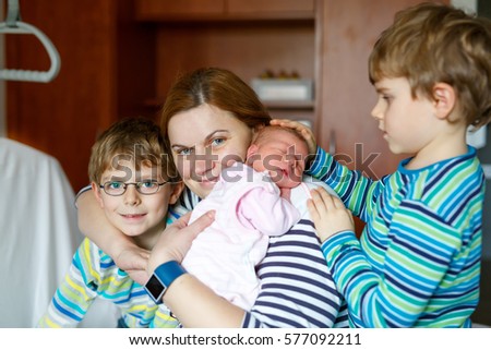 Mother holding newborn baby girl on arm. Two kids boys sitting by mum and sister. Siblings, love. Family on bed in hospital. New born child sleeping. Kids bonding. Mama of three children.