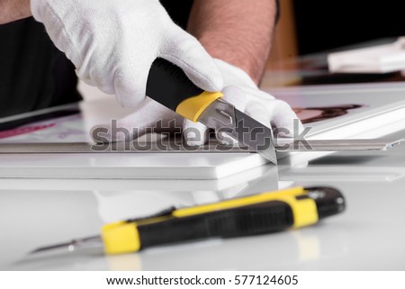 Professional hands in gloves cutting off edges of printed photography on white Foamex PVC panel board with another utility knife on table in foreground Royalty-Free Stock Photo #577124605