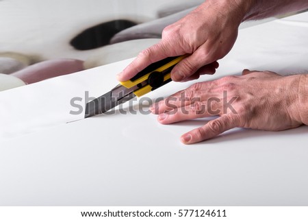 Designer male hands close-up shot making cuts of white wide sheet of paper using professional utility knife cutter Royalty-Free Stock Photo #577124611