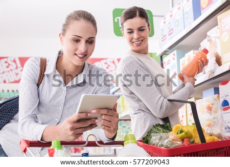 Happy female friends shopping together at the supermarket, they are connecting with a digital tablet, searching products and offers