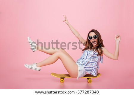 Cheerful young woman in sunglasses sitting on skateboard and having fun over pink background Royalty-Free Stock Photo #577287040