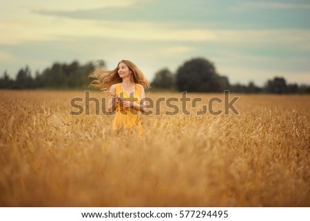 young girl with blond long hair with a smile on her face wearing a yellow sundress in the hands holding a bunch of grass the wind develops her hair standing in field or meadow in summer or autumn time