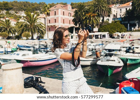 young beautiful woman traveling in Mediterranean, taking pictures on digital photo camera, summer vacation, resort style, sunglasses, sunny, tropical, sightseeing in port