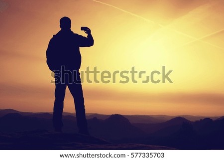 Tall hiker hold in one hand mobile phone and  take photo of misty landscape. Smart phone photography on  the peak of mountain at sunrise.