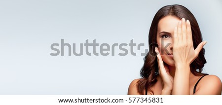 Smiling young woman, with eye, clossed by hand, covering part of her face, over bright grey background, with blank copyspace area for slogan or text message.  Royalty-Free Stock Photo #577345831