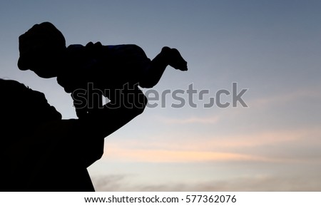 Silhouette of long hair girl raises her boy in the air with evening sky background.