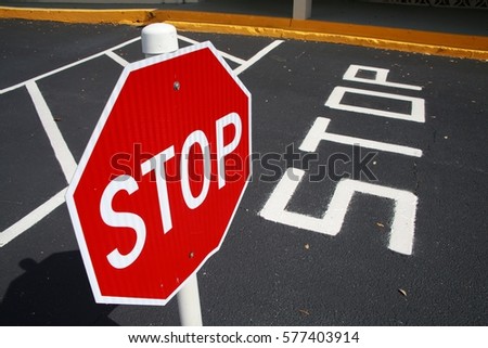 Red and White Stop Sign and Indication Painted on Black Pavement in Parking Lot with Yellow Curb in Background, Angle Facing Right, Bright Sunny Day in February