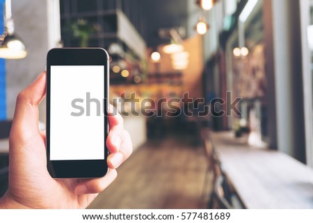 Mockup image of hand holding black mobile phone with blank white screen in modern cafe