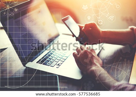 Male hands holding contemporary smart phone while sitting at desktop with laptop computer. Business man updating applications on cellular. Diagrams and statistics indicators with infographics effect