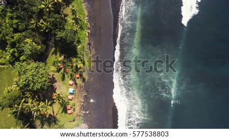 Top view aerial drone photo of black volcanic sand shore backed by thick palm trees tropical rainforest. Best camp on the beach where Indian ocean waves meet with coastline. Travel blog background
