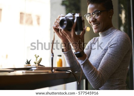 Portrait of cheerful female travel blogger taking picture via vintage camera while sitting in modern interior cafe enjoying trip planning to use photos in articles sharing impression with followers