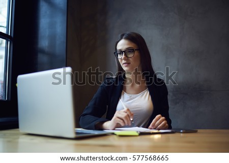 Young talented female journalist in trendy glasses watching online translation of political meeting ready to write article for prime time broadcast using wireless internet and laptop in newsroom
