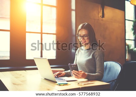 Experienced female editor in trendy eyewear of online issue coordinating work of content managers using laptop and wifi while reading notification on smartphone from bank confirming remittance 