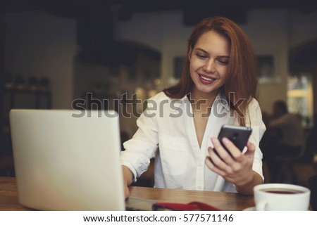 Attractive female international student testing new software for digital devices connecting smartphone and laptop to upgrade system while resting in cafe after lesson using free internet access 