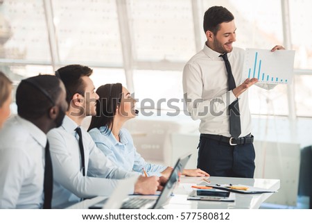 Attractive business people in suits and headsets are smiling and listening to their colleague while working with laptops in office