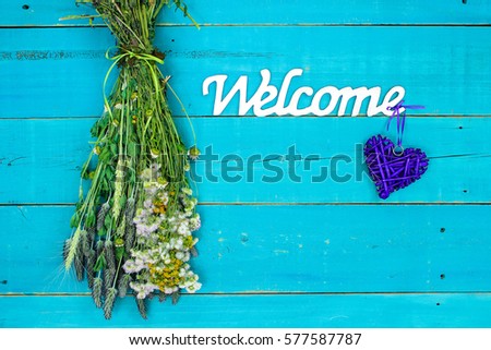 Welcome sign with heart and bouquet of dried meadow flowers hanging by rope on antique rustic teal blue wooden background; Mothers Day, home and love concept background with painted copy space