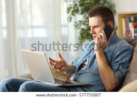 Angry man having problems on line with a laptop talking on the mobile phone with support service sitting on a couch in the living room at home Royalty-Free Stock Photo #577755007
