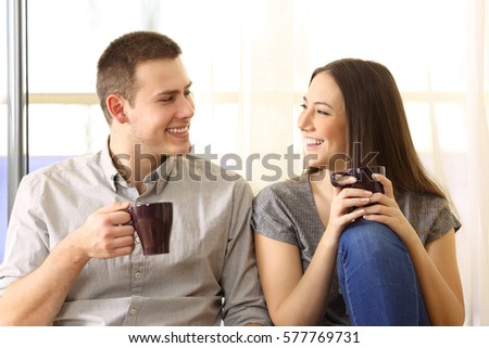 Front view of a happy couple talking and drinking coffee sitting on the floor near a window at home