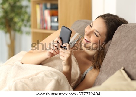 Happy girl using a smart phone covered with a blanket sitting on a comfortable couch in the living room at home