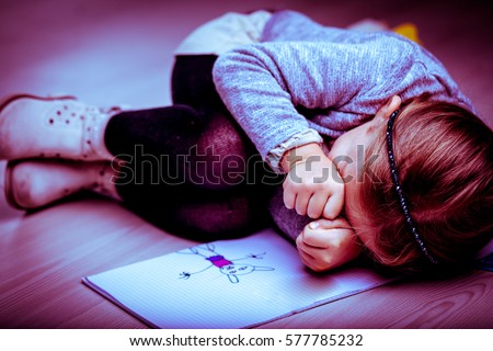 Upset little girl curled up on the bed next to her drawing rubbing her eyes with her fists as though crying, side vignette