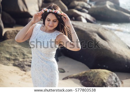 Young beautiful girl in a white dress at the sea beach with big stones. A wreath of flowers in her hair. Beauty portrait of female