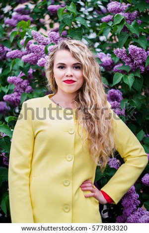 Portrait of blonde girl with wavy hair. Wearing yellow coat. Standing against blooming violet lilac tree. Walking outdoor at park. Springtime.
