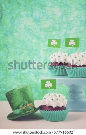 Happy St Patricks Day cupcakes with shamrock flags and green leprechaun hat against a green background on a white wood table, with applied retro style faded filters.