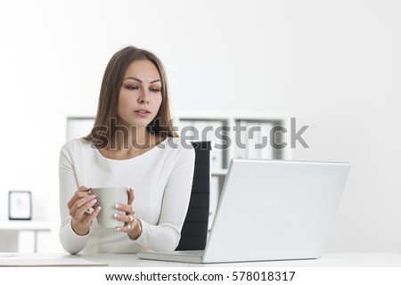 Pensive girl with a gray cup of coffee in a white office looking at her laptop screen. There are shelves with folders in the background.