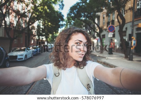 Cheerful curly woman taking a selfie photo on the street using her mobile phone, walking in a city