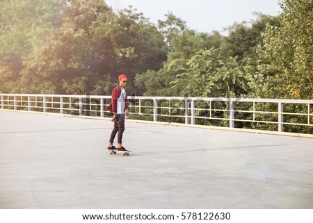 Picture of young dark skinned boy wearing sunglasses and cap skateboarding. Against the nature background.