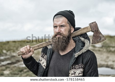 Handsome man hipster or guy with beard and moustache on serious face in hat and jacket holds rusty axe with wooden hilt outdoor on mountain top against cloudy sky on natural background, copy space Royalty-Free Stock Photo #578158465