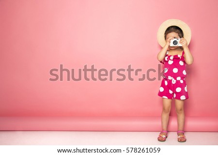 Portrait of cute little girl in straw hat and pink jumpers taking picture with white camera in the studio on pink background. Photography concept. Copy space for text