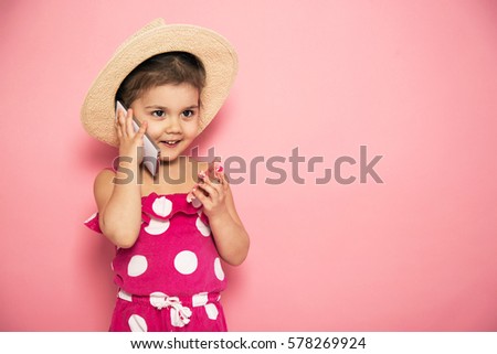 Portrait of cute little girl in pink jumpers and straw hat talking on the smartphone in the studio on pink background. Summer accessories. Communication concept.