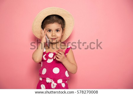 Portrait of cute little girl in pink jumpers and straw hat talking on the smartphone in the studio on pink background. Summer accessories. Communication concept.