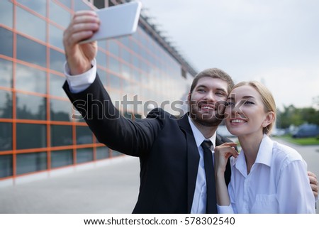 Young business partners standing in front of modern office, making selfie with mobile phone and smiling. Business people having coffee break outdoors