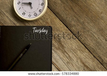 Business concept. Top view of  table clock, pen and notebook written with TUESDAY on wooden background. Copy space. Flat lay.