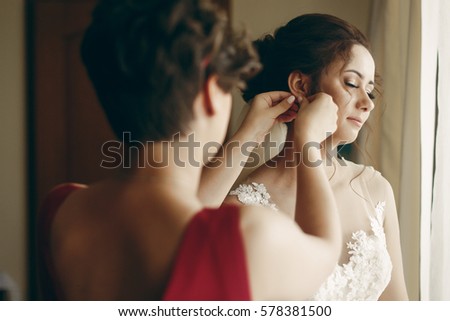 Beautiful bridesmaid helping brunette bride in white wedding dress put on luxury earrings, morning wedding preparation, bride with earrings close-up