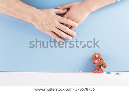 Man's hand is on top of a woman's hand. Nearby is a toy bear with the inscription "I love you." Blue background.