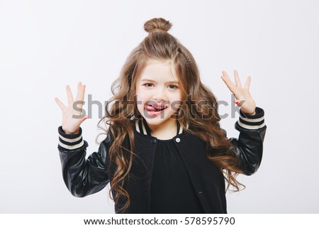 Portrait of little hipster girl in bomber jacket showing tongue. Posing. Curly modern hairstyle. Smiling. Studio