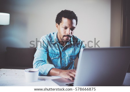Closeup view of pensive bearded African man using laptop at coworking studio on the wooden table.Concept of young people work mobile devices.Blurred background
