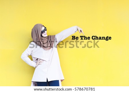 Asian young lady pointing her finger to the words typed Be The Change isolated with yellow background. Conceptual image for business, finance, inspiration and creativity.