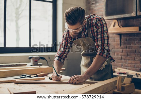 Bearded handsome cabinetmaker at the tabletop with pencil drawing sign on plank.  Stylish craftsman with brutal hairstyle and saved glasses work at his workstation. Royalty-Free Stock Photo #578729914