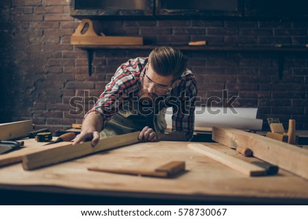 Bearded handsome cabinetmaker at the tabletop with tools.  Stylish craftsman with brutal hairstyle and saved glasses holding woodenplank at his workstation. Royalty-Free Stock Photo #578730067