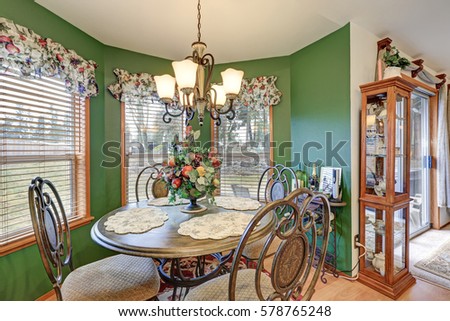 Amazing bright green breakfast nook boasts wrought iron table set lit by elegant chandelier, windows dressed in colorful curtains, small glass cupboard by the doors to backyard. Northwest, USA