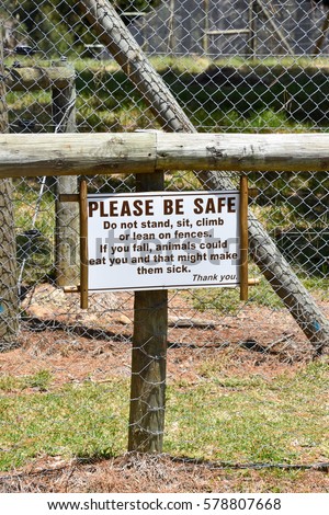A sign saying please be safe and do not stand sit climb or lean on fences, if you fall animals could eat you and that might make them sick