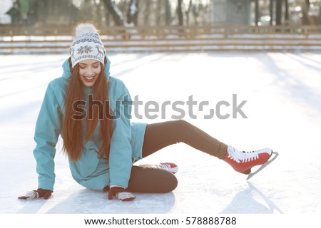 Pretty young girl in warm clothes and skates spending time at snowy  winter park while sitting on skating  rink after falling and laughing