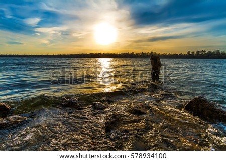 Sunset at Lake Jacomo just outside of Kansas City, Missouri.  it is shot horizontal with waves going over a rock and around a stump in the water