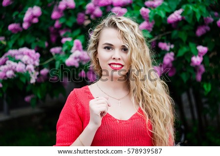 Portrait of pretty blonde girl with wavy hair, posing and looking and camera. Emotionally smiling. Wearing red blouse. Standing against blooming violet lilac tree. Walking outdoor at park. Springtime.