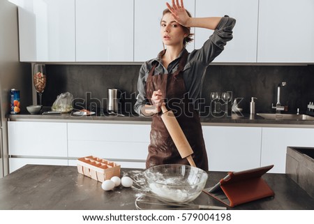 Photo of young woman standing in kitchen and cooking the dough. Looking at camera.